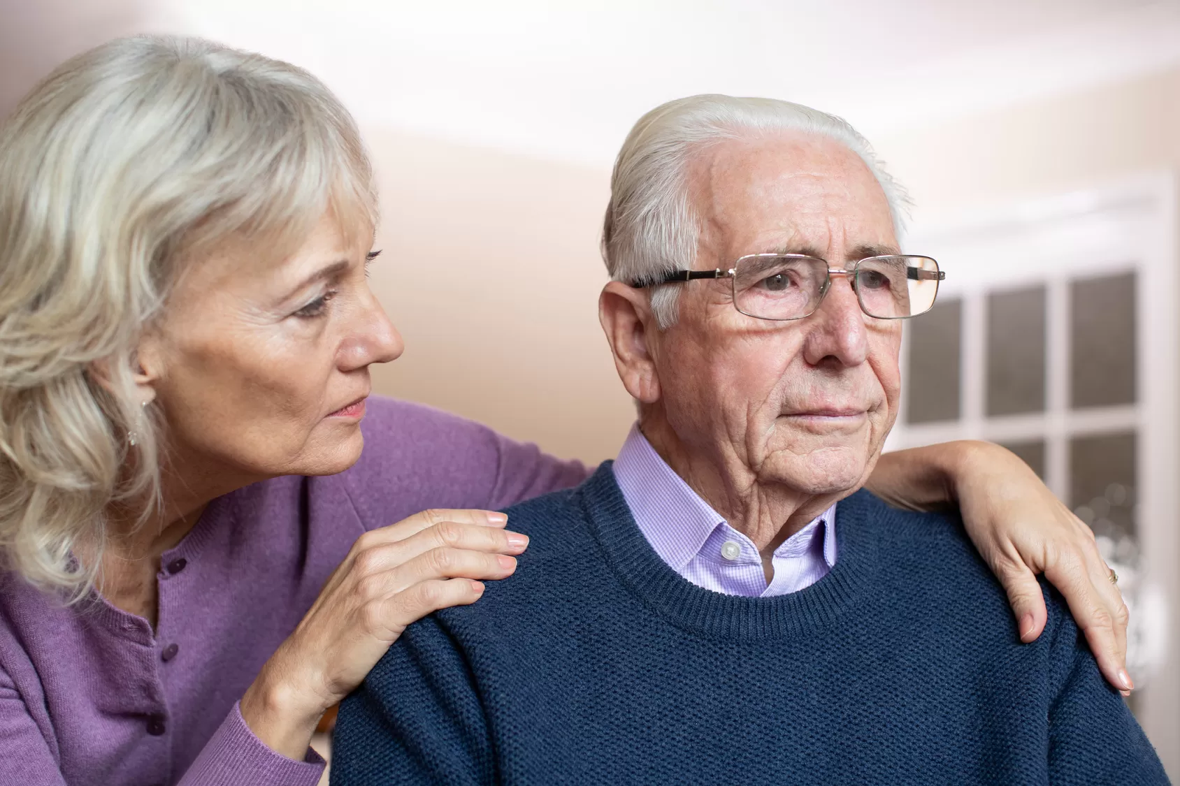 Elderly couple sitting together. Woman has her arms around a worried senior man